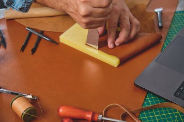 Bir zanaatkar, küçük bir atölyede, aletler ve dizüstü bilgisayarla çevrili bir tezgahta kahverengi deriyi kesmek için hassas bir bıçak kullanır.