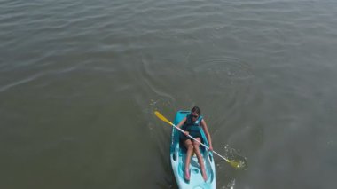 Bir kız bir su kütlesinin içinde kanoyla kürek çekiyor. Su sakin ve gökyüzü berrak.