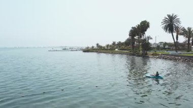 Bir kız rıhtımın yanındaki okyanusta kano yapıyor. Su sakin ve gökyüzü kapalı.