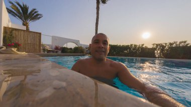 Bir adam arka planda güneş batarken havuzda yüzüyor. Gülümsüyor ve anın tadını çıkarıyor.