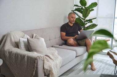 Evden çalışan, dizüstü bilgisayarlı rahat bir koltukta oturan, dairesinin rahatlığının keyfini çıkaran bir adam.