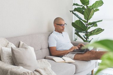 Evden çalışan bir adam, rahat oturma odasındaki kanepede dizüstü bilgisayar kullanıyor.