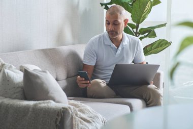 Adam evde çalışıyor, rahat bir koltukta otururken dizüstü bilgisayar ve akıllı telefon kullanıyor.