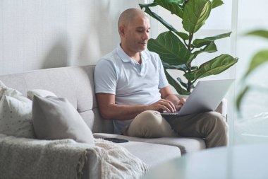 Evden çalışan, dizüstü bilgisayarlı rahat bir koltukta oturan, dairesinin rahatlığının keyfini çıkaran bir adam.