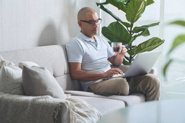 Adam evde çalışıyor, kanepede dizüstü bilgisayarını kullanırken bir fincan kahvenin tadını çıkarıyor.