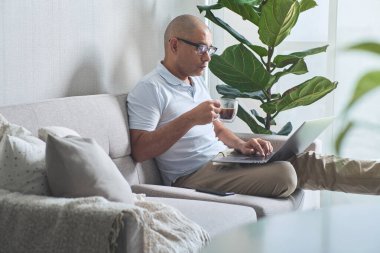 Gülümseyen serbest yazar evde çalışmaktan zevk alıyor, kanepede dizüstü bilgisayar ve kahveyle dinleniyor.