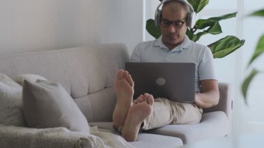 Evden çalışan bir adam kanepede dizüstü bilgisayar ve kulaklık kullanarak oturuyor.