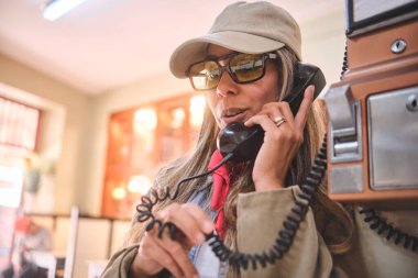 Kadın ankesörlü telefon kulübesinde telefon ediyor, güneş gözlüğü ve şapka takıyor.