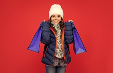 Şişkin ceketli ve şapkalı mutlu çocuk. Kırmızı arka plandan alışveriş yapan genç bir kız. Çocuk alışveriş torbalarını tutuyor. kış satışları.