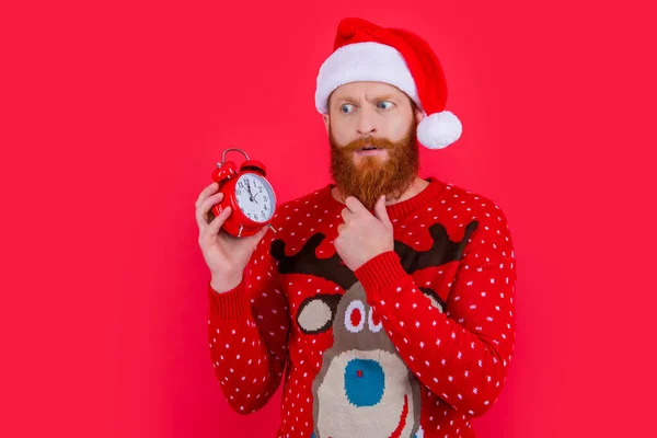 Puzzled man in Santa Claus hat stroking beard looking at alarm clock, time. New Year countdown. Time to celebrate. Christmas time. Merry Christmas.