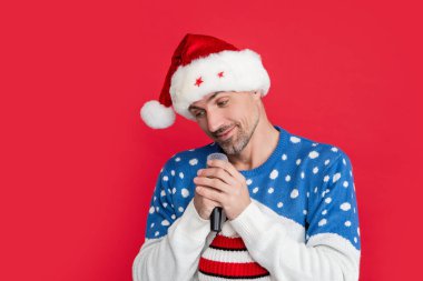 Yılbaşı şarkısı. Utangaç adam stüdyoda mikrofonla Noel şarkısı söylüyor. Adam kırmızı arka planda izole bir şekilde noel şarkısı söylüyor..