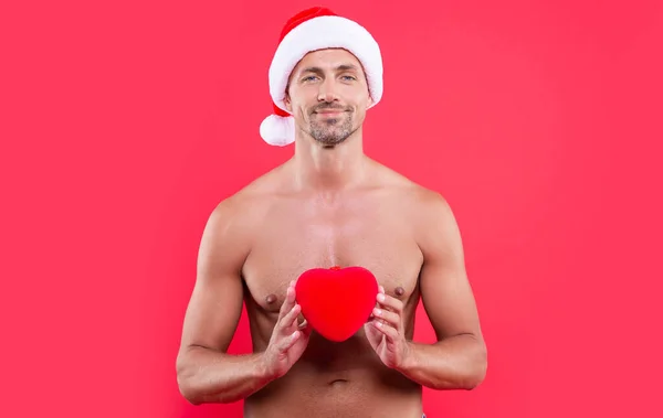 happy man hold heart at christmas in studio. christmas heart and man with torso. shirtless man in christmas santa claus hat with heart gift. christmas man isolated on red background.