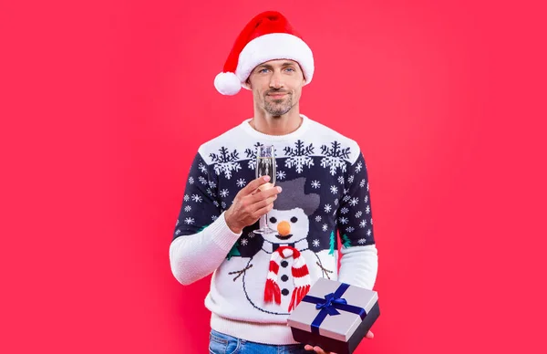 cheerful christmas man isolated on red background. man hold gift for christmas in studio. christmas gift and man with champagne. man in christmas santa hat with gift and champagne.