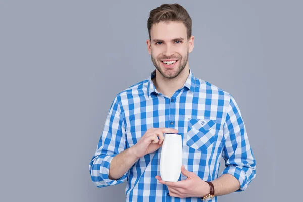 happy man with unbranded cosmetic in studio. unbranded cosmetic for man. stylish man hold bottle of unbranded cosmetic. man in checkered shirt hold unbranded cosmetics isolated on grey background.