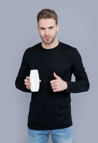 thumb up. unbranded cosmetic for man. handsome man hold bottle of unbranded cosmetic. man hold unbranded cosmetics isolated on grey background. man with unbranded cosmetic in studio.