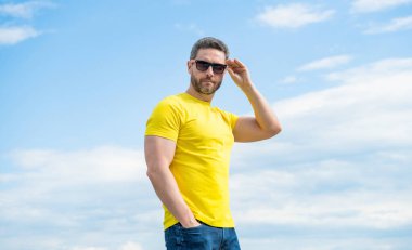 Gökyüzü arka planında sarı gömlekli ve gözlüklü bir adam..