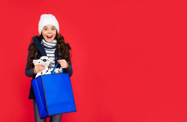 穿着宽松夹克 头戴帽子的孩子很惊讶 在红色背景下购物后的少女 带有复制空间 小孩拿着购物袋和礼物玩具 冬季销售 玩具店 — 图库照片