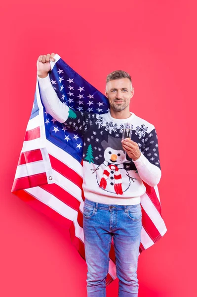 man hold usa flag and champagne in christmas studio. man in christmas sweater holding usa flag. man in christmas sweater with usa flag. christmas man isolated on red background.