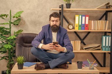 Kafası karışmış profesyonel bir adam akıllı telefondan mesaj okuyor. Ofis masasında bağdaş kurmuş oturuyor..