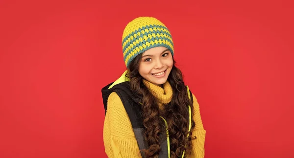 express positive emotion. winter fashion. positive kid with curly hair in hat. teen girl on red background. portrait of child wearing warm clothes.