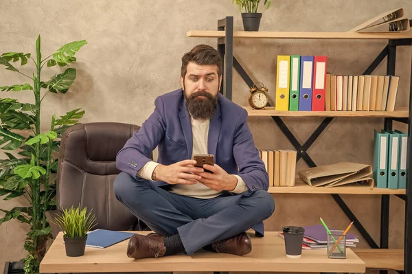 Professional man with puzzled look reading message in smartphone sitting cross-legged on office desk, sms.