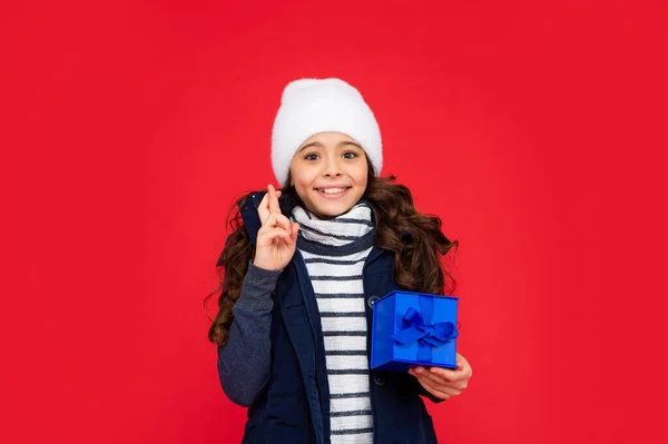 happy child in puffer jacket and hat hold box. kid with present. teen girl on red background. winter holidays. crossing fingers to make a wish.