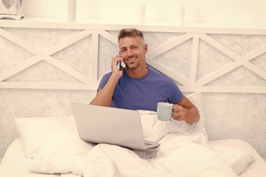 Telefonda sohbet ediyorlar. Mutlu adam cep telefonunda dizüstü bilgisayarla konuşuyor. Çevrimiçi sohbet. Yatakta sabah kahvesi.