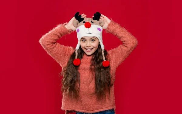glad teen girl in winter fashion hat isolated on red. teen girl wear sweater and winter fashion hat in studio. winter fashion for teen girl. teen girl fashion model in winter earflap hat.