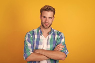 Ciddi yakışıklı bir genç portresi. Kollarını işaretli tişörtün içinde tutuyor. Sarı arka plan, kendinden emin..