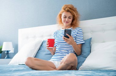 Telefonda internette kahve sörfü yapan gülümseyen bir kadın. Evde telefonuyla internette sörf yapan bir kadın. Kadının evinde internet sörfü.