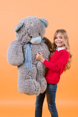 Hayvan bebek, mevcut olan çocuk ve hediye. Mutlu çocukluk kavramı. Tatil, doğum günü, yıldönümü kutlamaları. Kız büyük oyuncak ayı turuncu arka plan üzerinde tutun. Çocuk gülümseme ile gri yumuşak oyuncak.