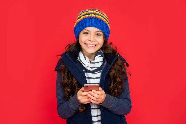 express positive emotion. greeting with winter holidays. positive kid chatting online in hat on red background. teen girl in down vest texting on smartphone. portrait of child blogging with phone.