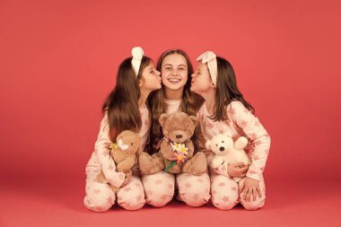 Pijamalı mutlu kızlar ablamı öpüyor. Oyuncak ayılarla oturuyorlar. Pembe arka plan, kız kardeş sevgisi..