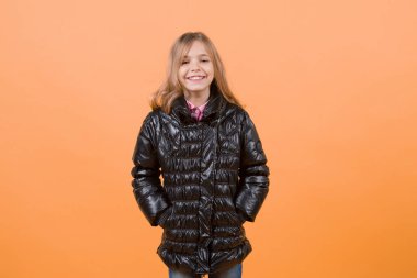 Turuncu arka plan üzerinde siyah palto kız. Moda, sonbahar bir tarz, trend. Uzun sarı saçlı çocuk modeli gülümseme. Çocuk güzellik, göz, saç modeli. Mutlu çocukluk kavramı, kopya alanı