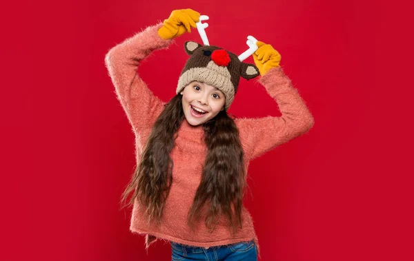 funny teen girl in winter fashion hat isolated on red. teen girl wear sweater and winter fashion hat in studio. winter fashion for teen girl. teen girl fashion model in winter earflap hat.