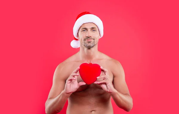 cheerful christmas man isolated on red background. man hold heart at christmas in studio. christmas heart and man with torso. shirtless man in christmas santa claus hat with heart gift.