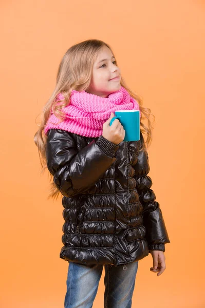 Girl with blue cup smile on orange background. Hot drink in cold weather. Child hold mug in black jacket and pink scarf. Tea or coffee break. Autumn season relax concept.
