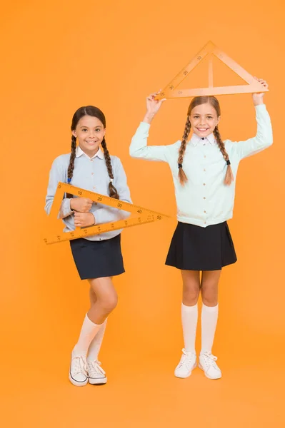 Measuring angles at geometry. Cute schoolgirls holding triangles for geometry lesson on yellow background. Little girls learning geometry. Small children with geometrical tools for geometry.