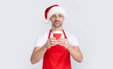 Kırmızı Noel Baba şapkalı ve önlüklü olgun bir adam elinde kahve fincanı tutuyor..