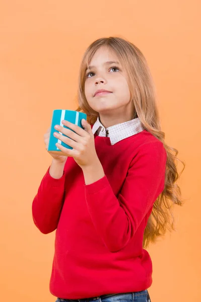 Health and healthy drink. Girl with long blond hair in red sweater with mug. Child with serious face hold blue cup on orange background. Tea or coffee break. Thirst, dehydration concept.