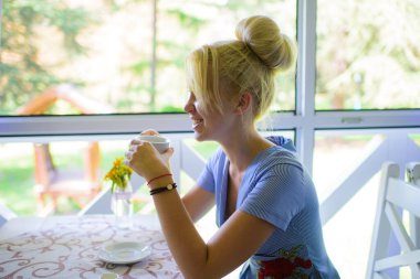 Kadın pencerenin yanında oturuyor, doğa arka planında. Kafe ve iletişim konsepti. Kafede ya da restoranda kahve ya da çay içen bir kız. Gülen suratlı kız arkadaş komik bir hikaye dinliyor..