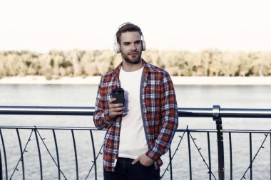 Kahve fincanı açık havada traşsız bir adam. Gündelik erkek hippi modası. Kulaklıklı adam, dinle. Müzik tarzı. Kahve içerken para kazanmak. Ses kalitesi. Günaydın içecek..