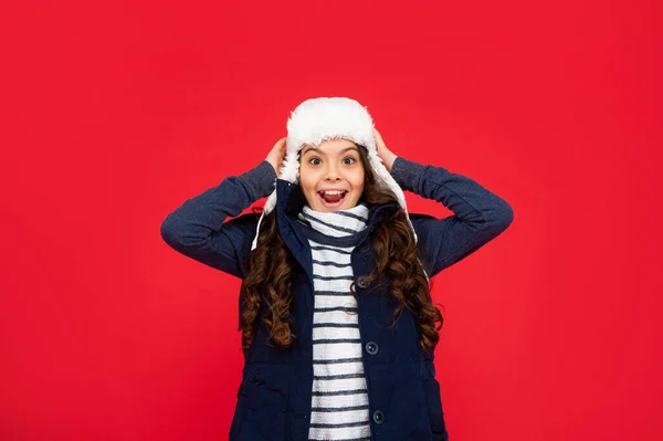 express positive emotion. winter fashion. happy kid with curly hair in earflap hat. teen girl on red background. portrait of child wearing warm clothes.