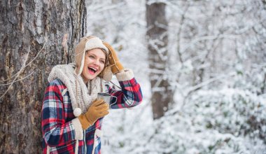 Eldivenli kadın ve şapkalı kadın karlı ormanda çay içiyor. Soğuk bir günde açık havada. seyahat ve yürüyüş. Yürüyüş yapan kız bir fincan sıcak çay içer. Kendini ısıt. Kadın kış içkisi. Kız kar yağışı seviyor.