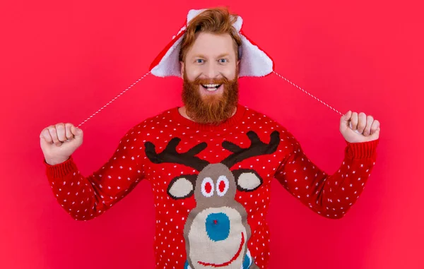 冬の帽子をかぶった幸せな男と赤い上に隔離されたXmasセーター クリスマスと新年のスタジオを祝う髭の男 新年会 明けましておめでとうございます — ストック写真