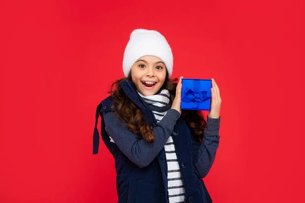 surprised child in puffer jacket and hat hold box. kid with present. teen girl on red background. winter holidays.