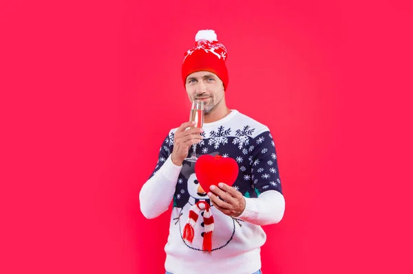 christmas man isolated on red background. man drink champagne for christmas in studio. christmas heart gift and man in sweater. man in christmas hat with champagne and heart.