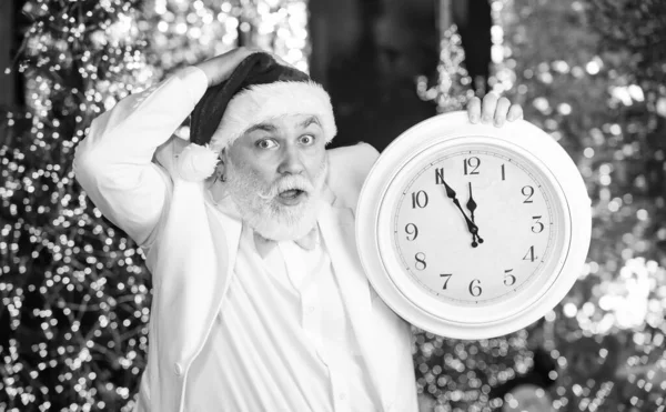 Senior santa man in tuxedo. Garland illumination background. Bearded businessman in santa hat. Christmas eve. Santa claus counting time. Midnight concept. New year countdown. Man with retro clock.