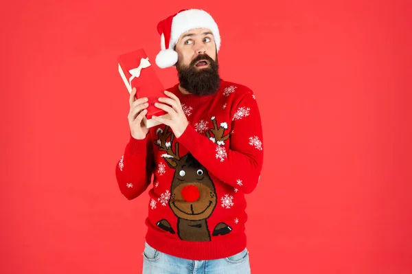 戴着圣塔克洛斯帽的满脸胡须的男人很惊讶 穿着冬季针织毛衣的野蛮嬉皮士 一个成熟的家伙 带着红色背景的节日礼品盒 拳击日圣诞节购物 新年快乐 快乐的圣诞节 — 图库照片