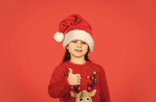 stock image Winter holidays sales. small girl santa claus hat red background. ready for new year party. christmas my favorite holiday. love winter holidays. in xmas mood. little santa helper show thumb up.